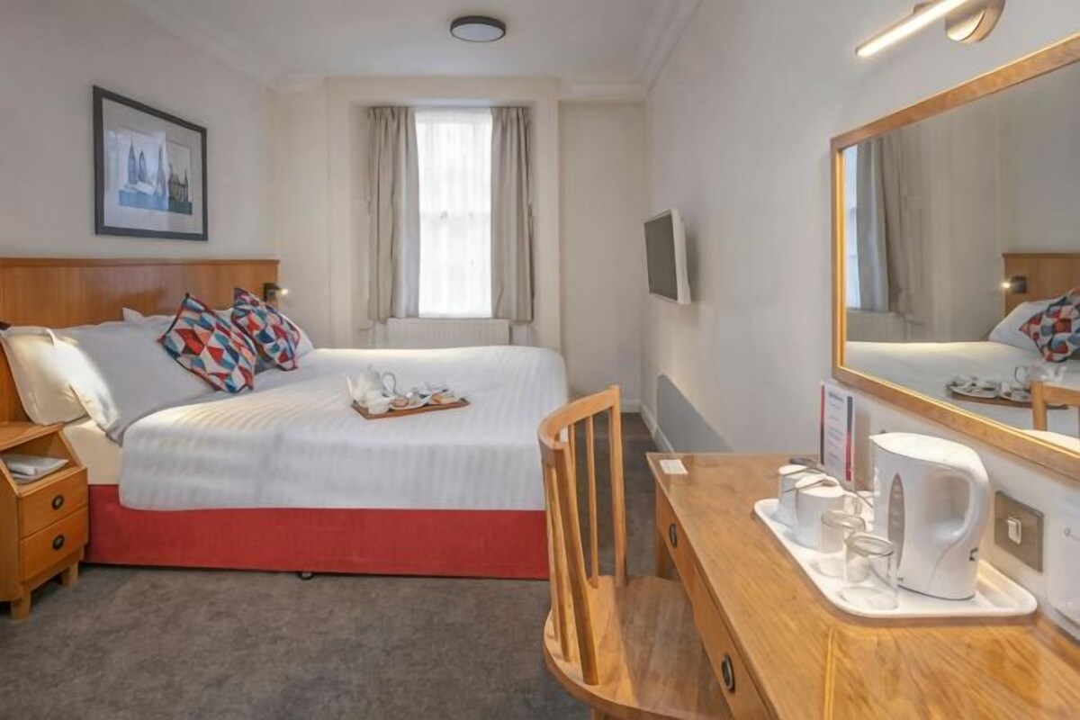 A hotel room with a bed, bedside table, chair, desk, mirror, window, and a framed picture on the wall.