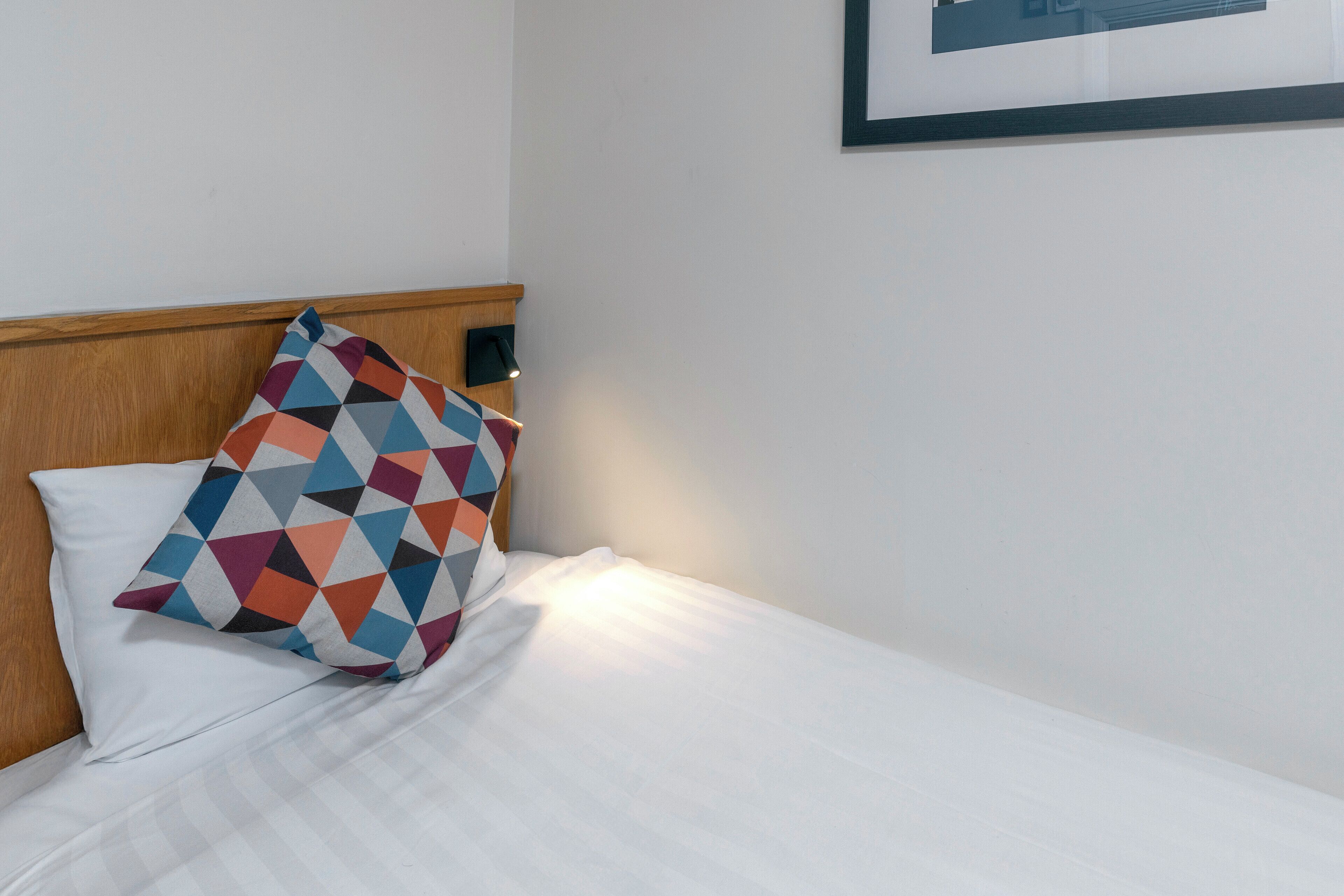 A neatly made bed with a colorful geometric pillow and a framed picture on the wall.