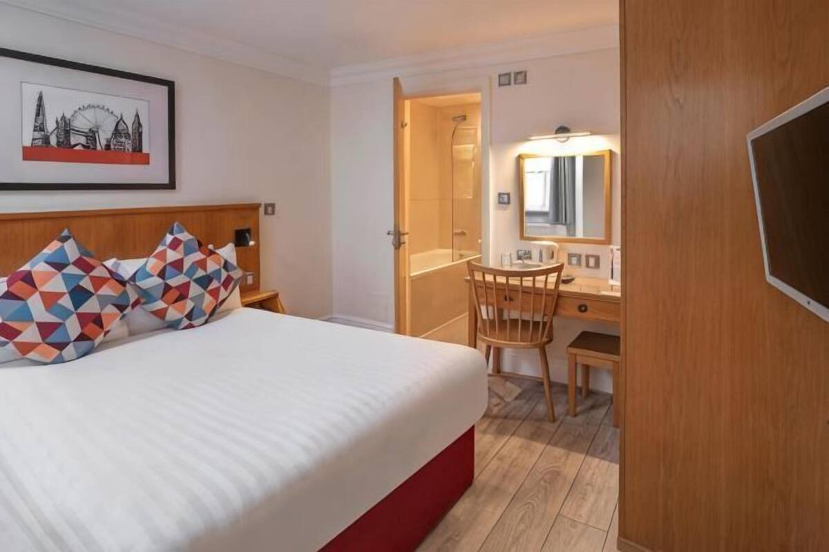 A hotel room with a bed, a desk, a chair, a mirror, and a framed picture on the wall.