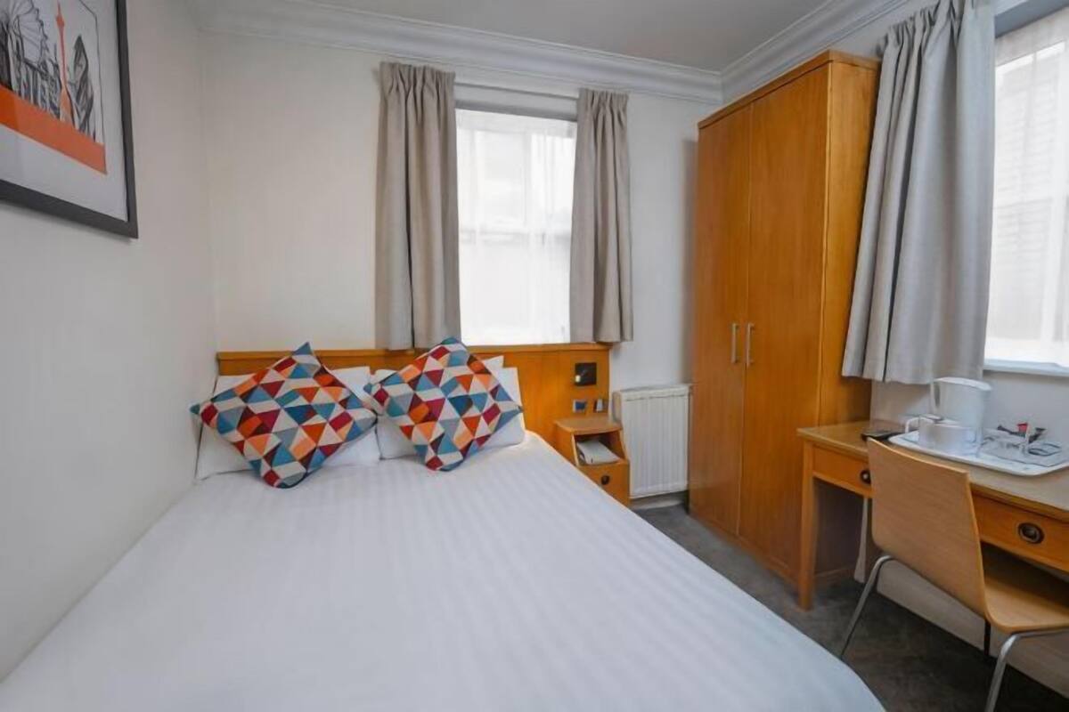 A hotel room with a bed, two pillows, a desk, a chair, a wardrobe, and a window with curtains.