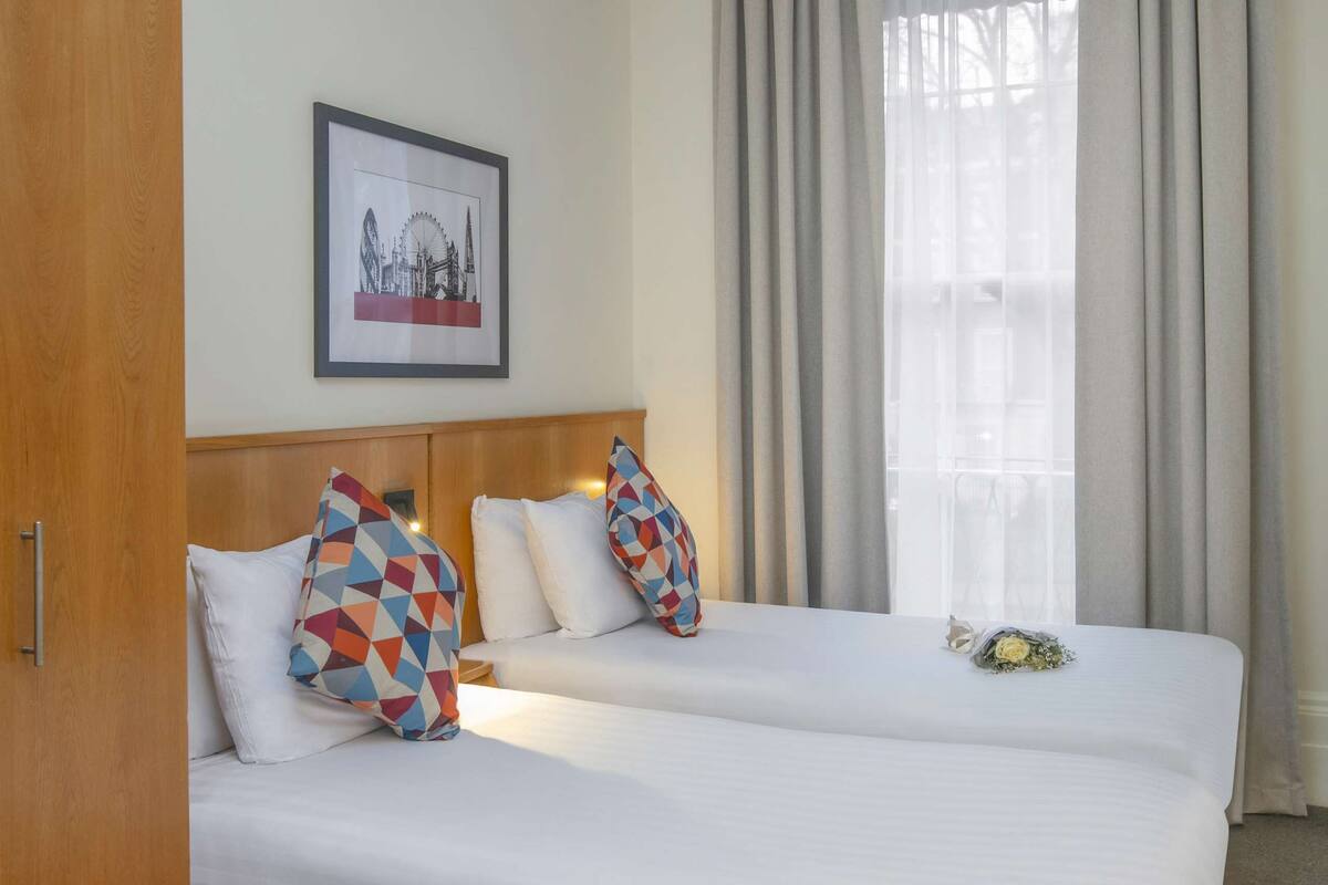 A hotel room with a double bed, a headboard, a framed picture on the wall, a window with curtains, and a wooden wardrobe.