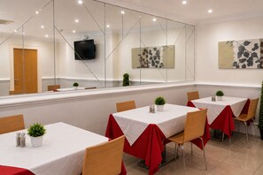 A restaurant with multiple tables, each with a white tablecloth and red table runner. There are potted plants on the tables, and the room has a modern design with a large mirror and artwork on the walls. - Queensway Hotel, Sure Hotel Collection by Best Western (London)