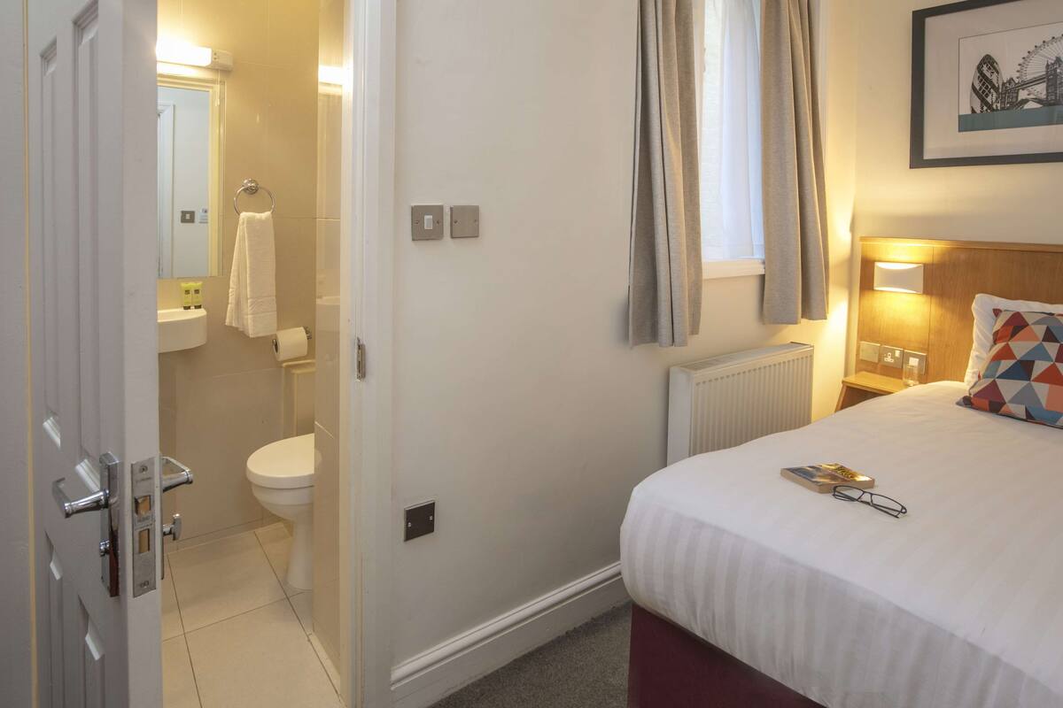 A hotel room with a bed, a bedside table with a lamp, a window with curtains, a bathroom with a toilet and sink, and a framed picture on the wall.