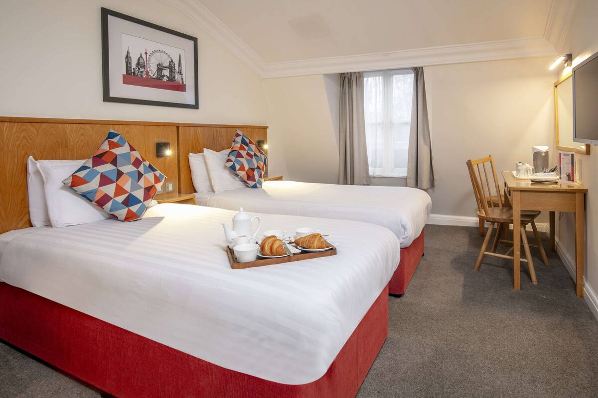 A hotel room with two beds, a desk with a chair, a framed picture on the wall, and a tray with breakfast items on the beds.