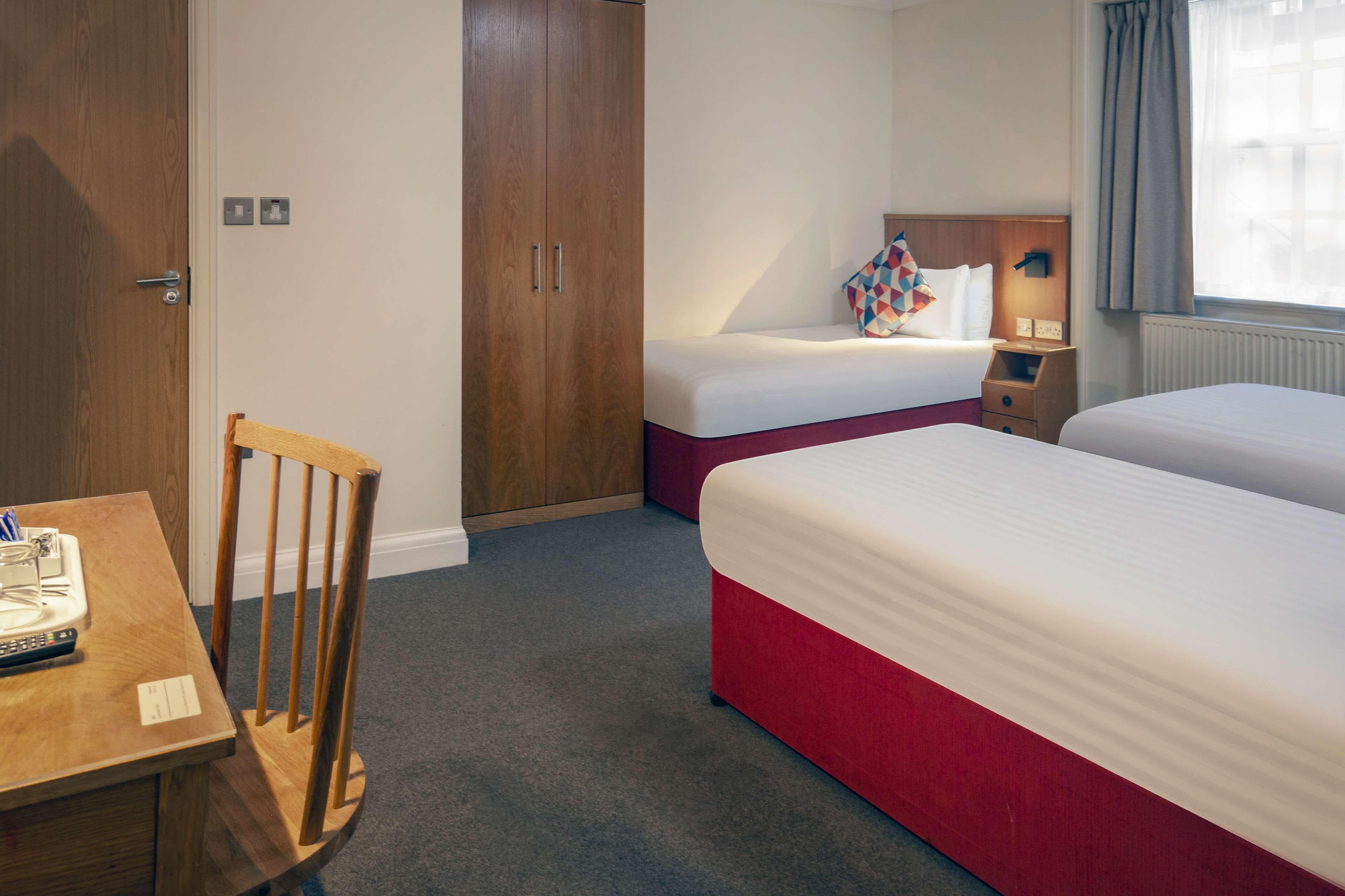 A hotel room with two beds, a wooden wardrobe, a desk with a chair, and a bedside table.