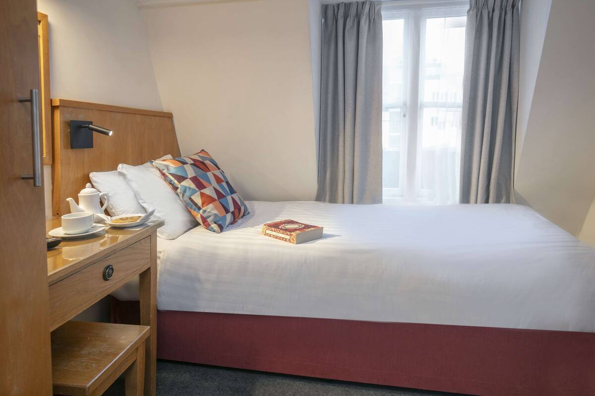 A neatly made bed with a white and red bedspread, a colorful pillow, and a book on the bed. There is a wooden nightstand with a cup and saucer, a kettle, and a remote control. A window with curtains is visible in the background.