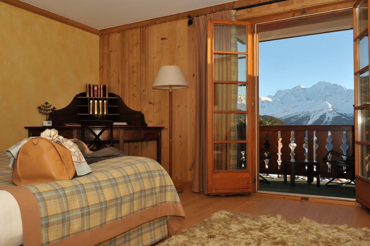 A bedroom with a bed, a wooden desk, a lamp, and a view of snow-capped mountains through a glass door.