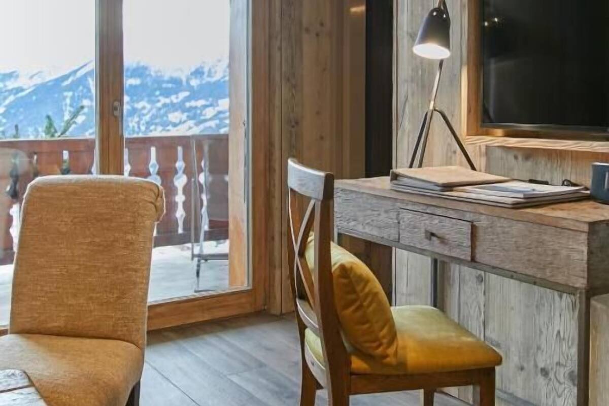 A room with a wooden desk, two chairs, a television, and a view of mountains through large windows.