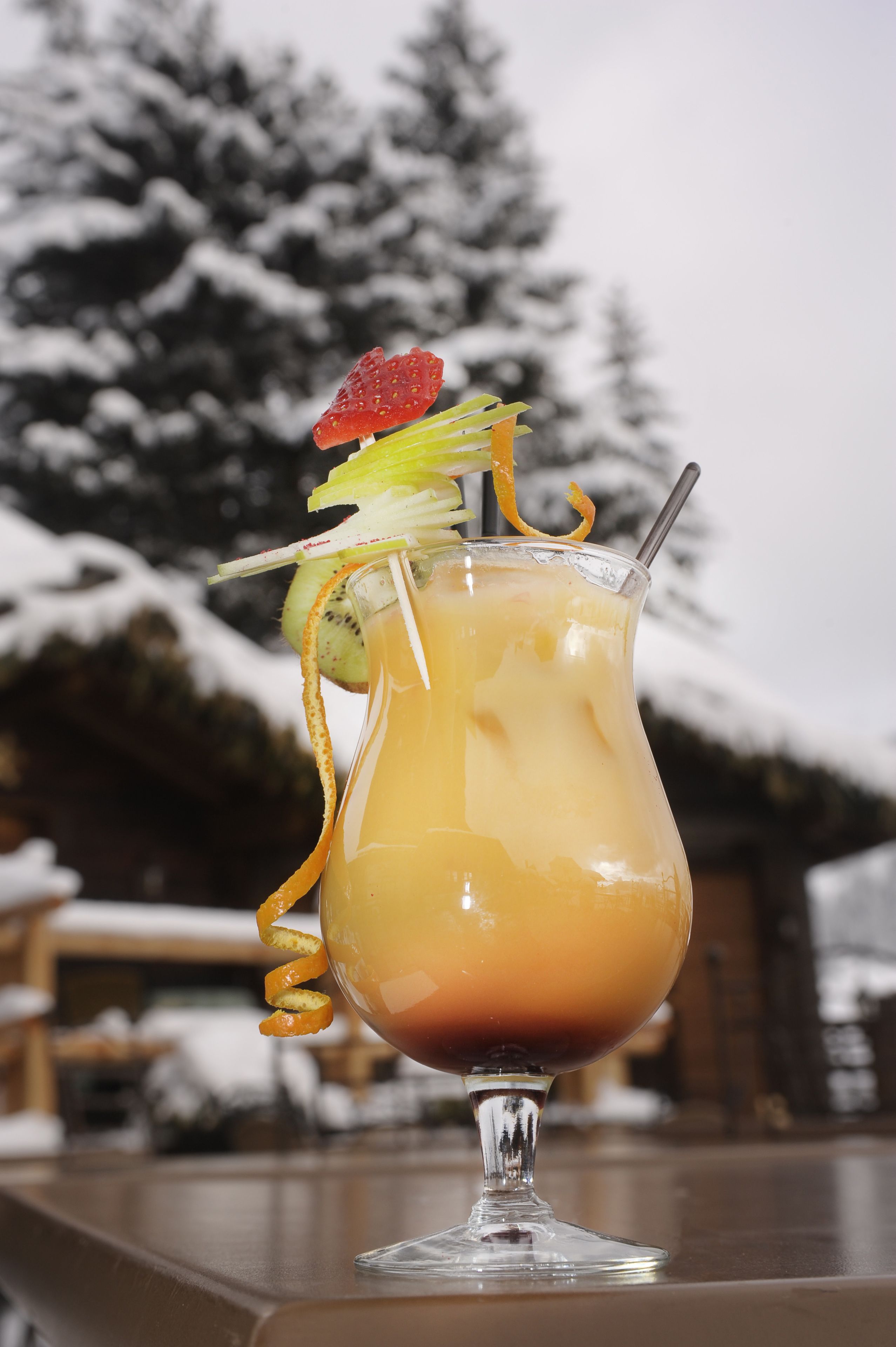 Un cocktail servi dans un verre, agrémenté d’une fraise et de tranches de kiwi, avec une paille, posé sur une table devant un décor enneigé.