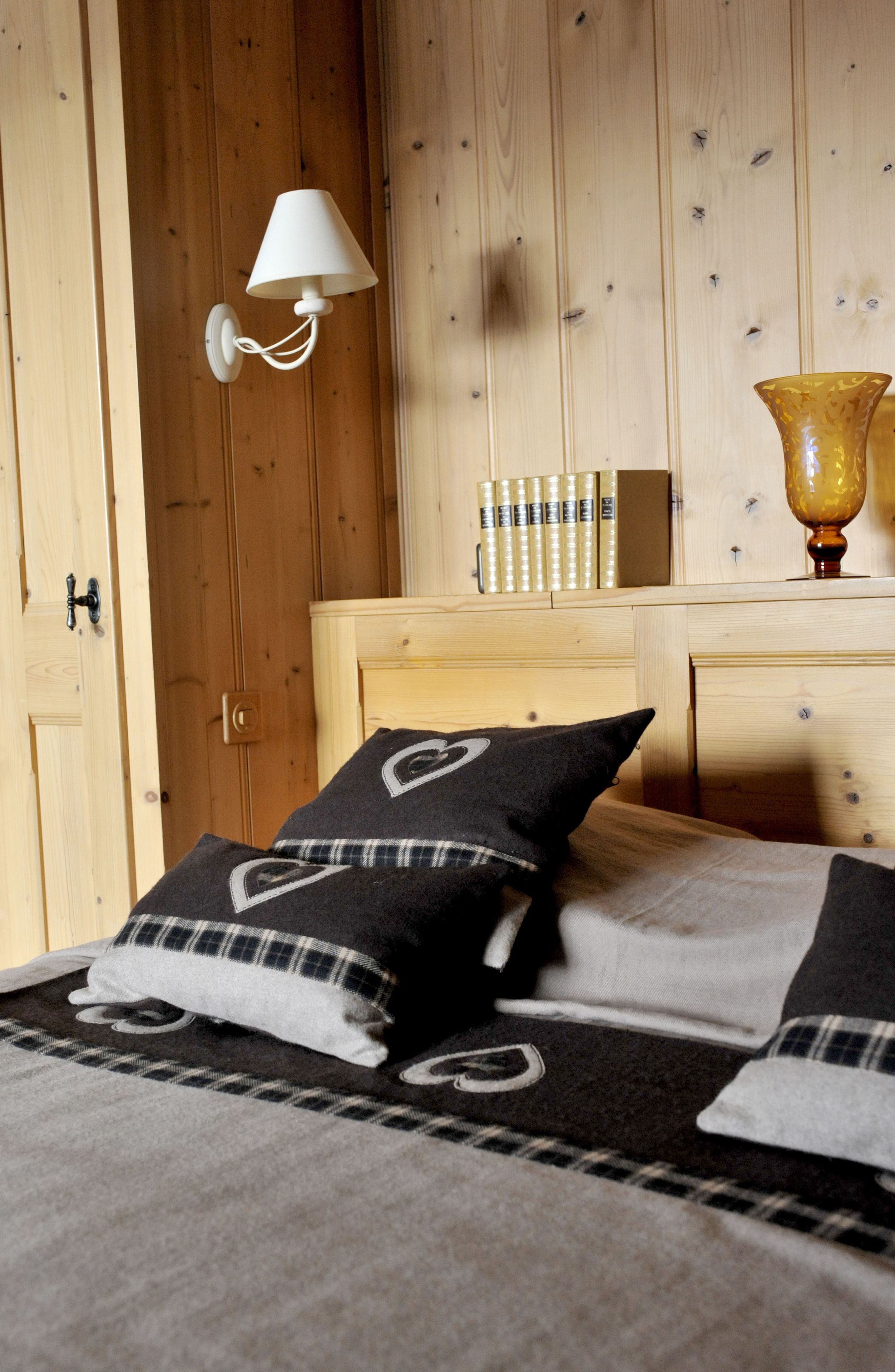 Ein Zimmer mit Holzwänden, einem Bett, einer Wandlampe, einem Nachttisch mit Büchern und einer Vase.