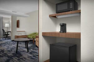 Habitación de hotel con kitchenette, que incluye microondas, cafetera y refrigerador. Hay un escritorio con una silla, una lámpara y un sillón verde. El piso está alfombrado y la pared es blanca con textura.