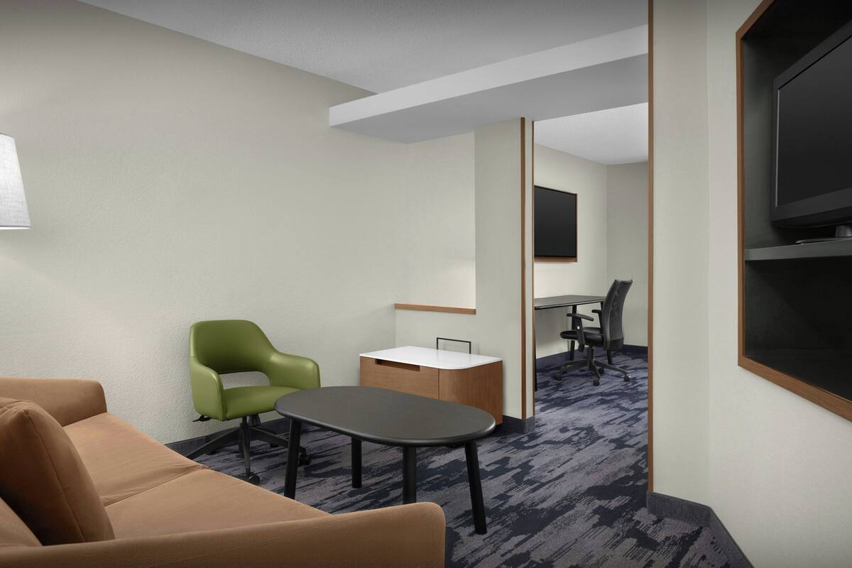 Une chambre d’hôtel avec un canapé marron, un fauteuil vert, une petite table, un bureau avec une chaise et un téléviseur fixé au mur.