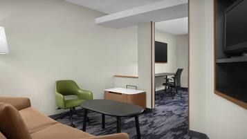 A hotel room with a brown sofa, a green chair, a small table, a desk with a chair, and a TV mounted on the wall.