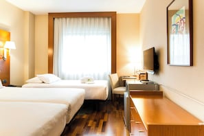 Habitación de hotel con dos camas, un escritorio, una silla, un televisor y un ventanal con cortinas.