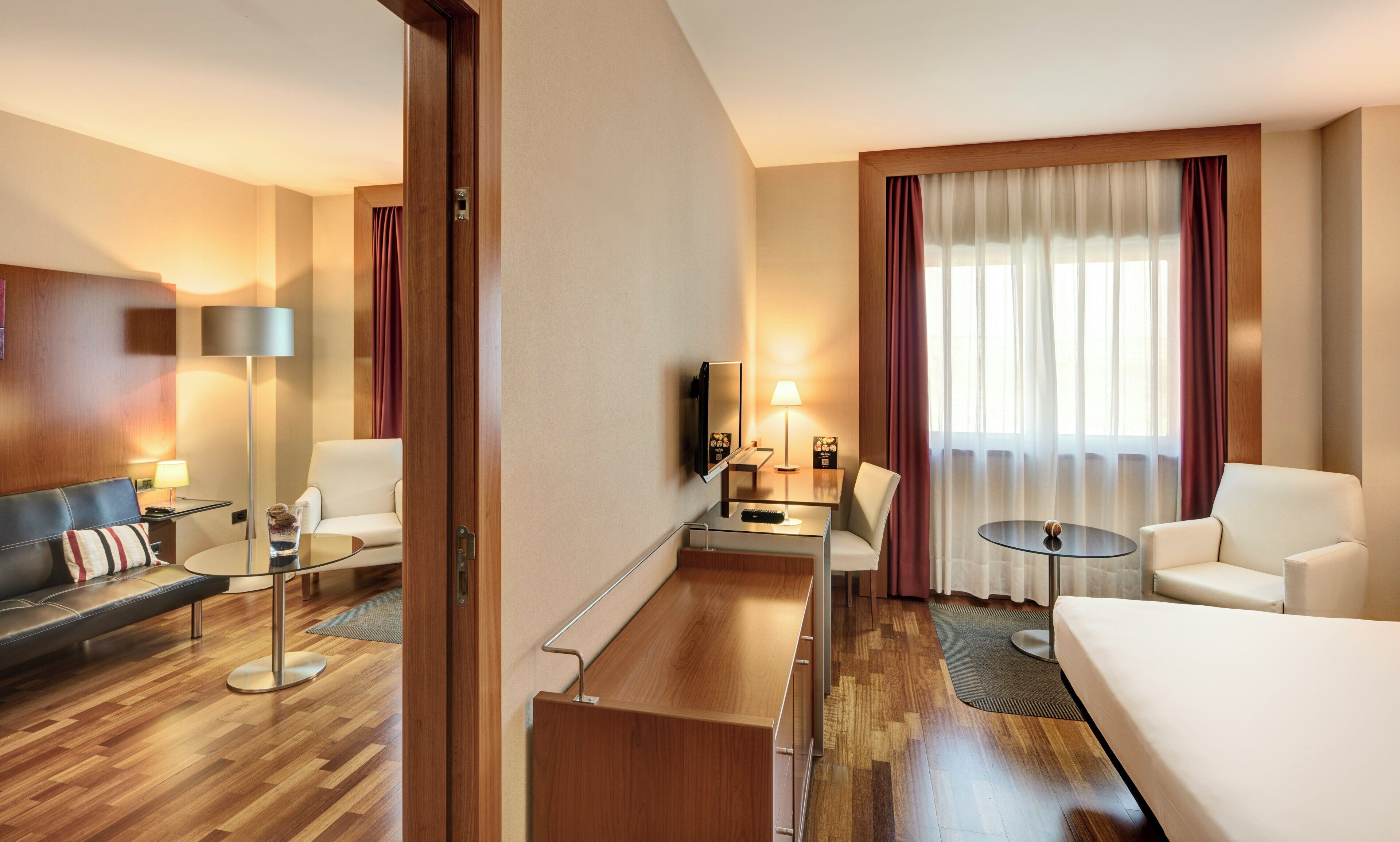 Habitación de hotel con sofá, sillones, una pequeña mesa, televisión y ventana con cortinas.
