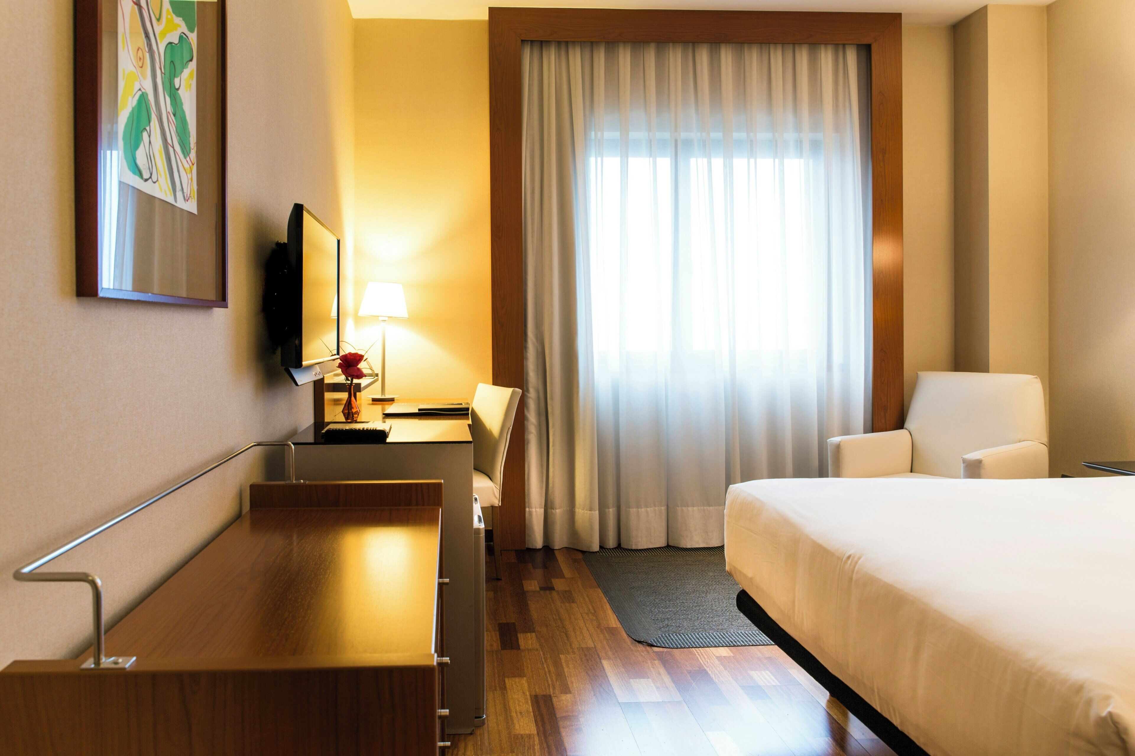 A hotel room with a bed, a desk with a lamp, a chair, a television, and a large window with curtains.
