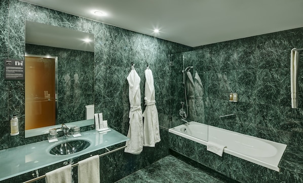 Un baño moderno con pared de mármol, lavamanos de vidrio, bañera y dos toallas blancas colgadas de ganchos.