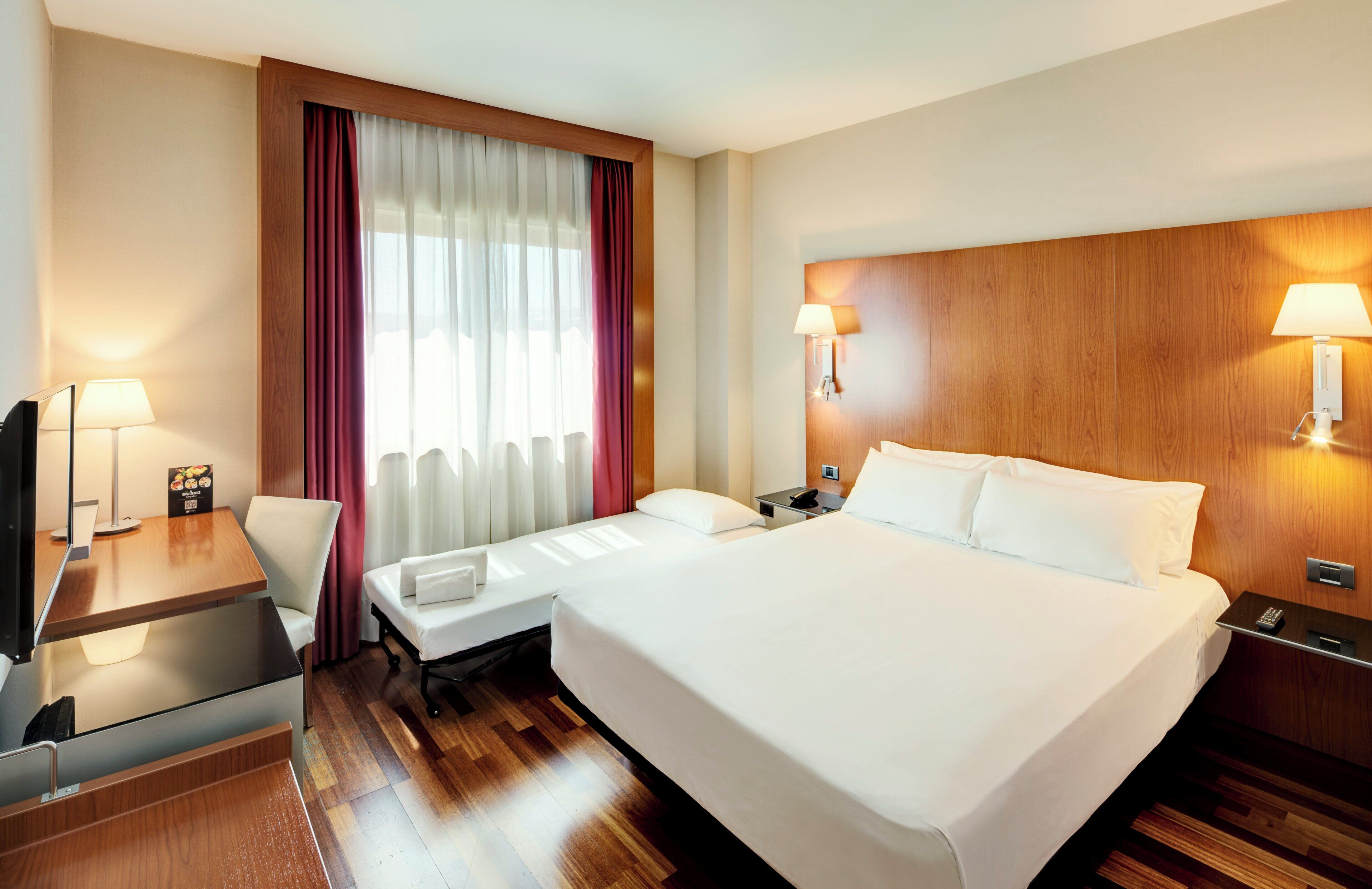 Habitación de hotel con cama, escritorio, silla, televisor, lámpara y ventana con cortinas.