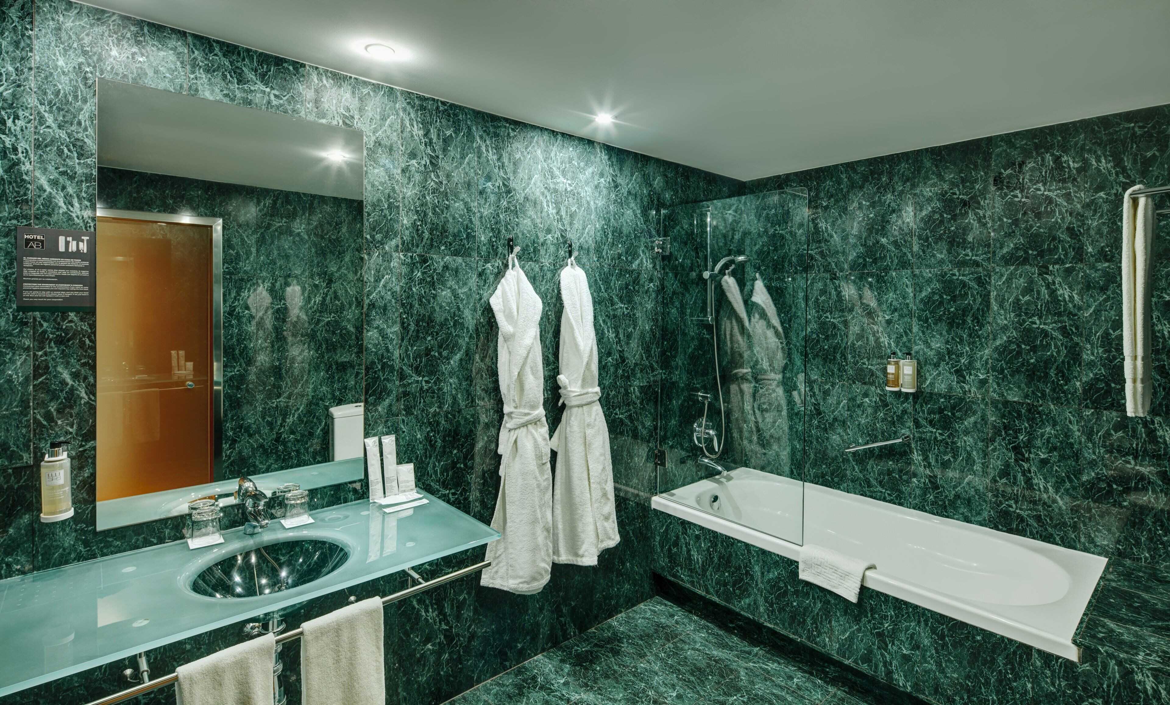 A modern bathroom with a glass sink, a large mirror, a bathtub, and marble walls.