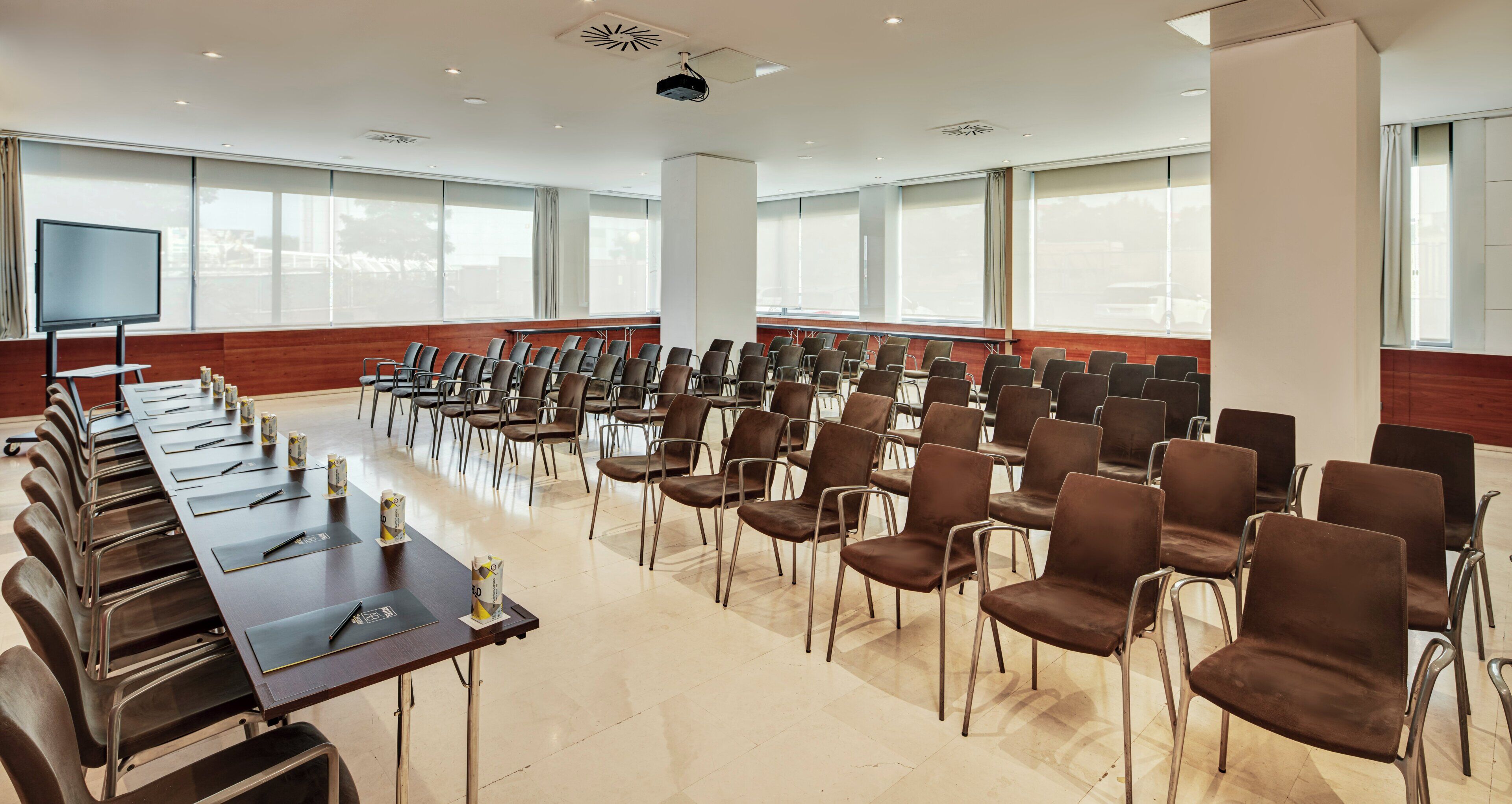 Un salón de conferencias con filas de sillas, una larga mesa con proyector y ventanas amplias.