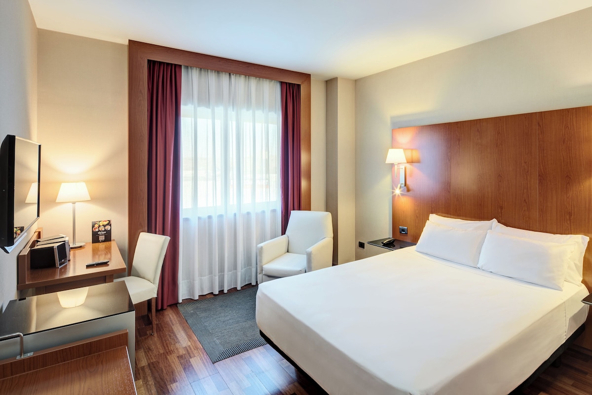 A hotel room with a large bed, a desk with a chair, a TV, and two bedside lamps.