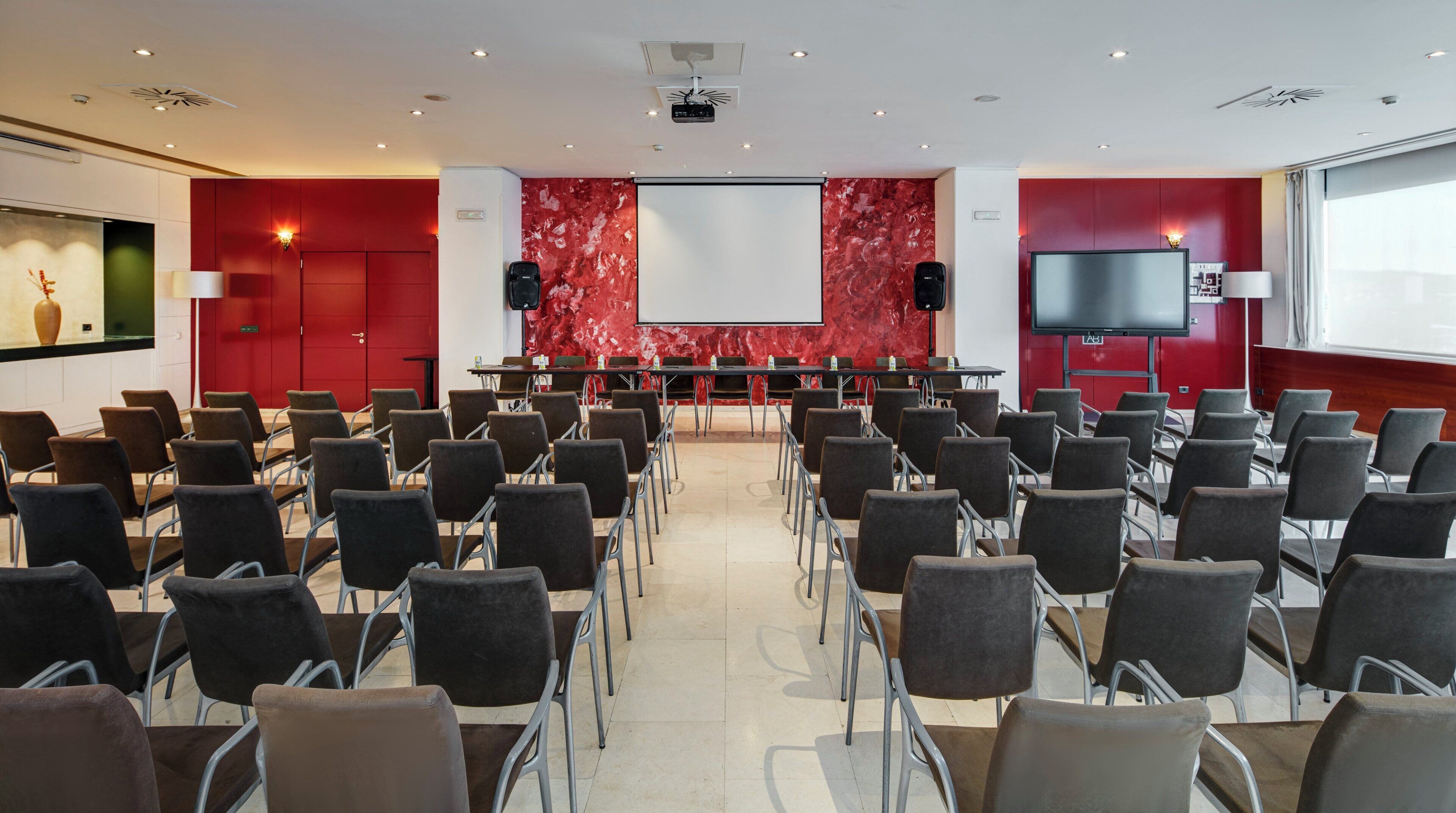 Une salle de conférence avec des rangées de chaises, un écran de projection, un grand téléviseur à écran plat et un mur en marbre rouge.