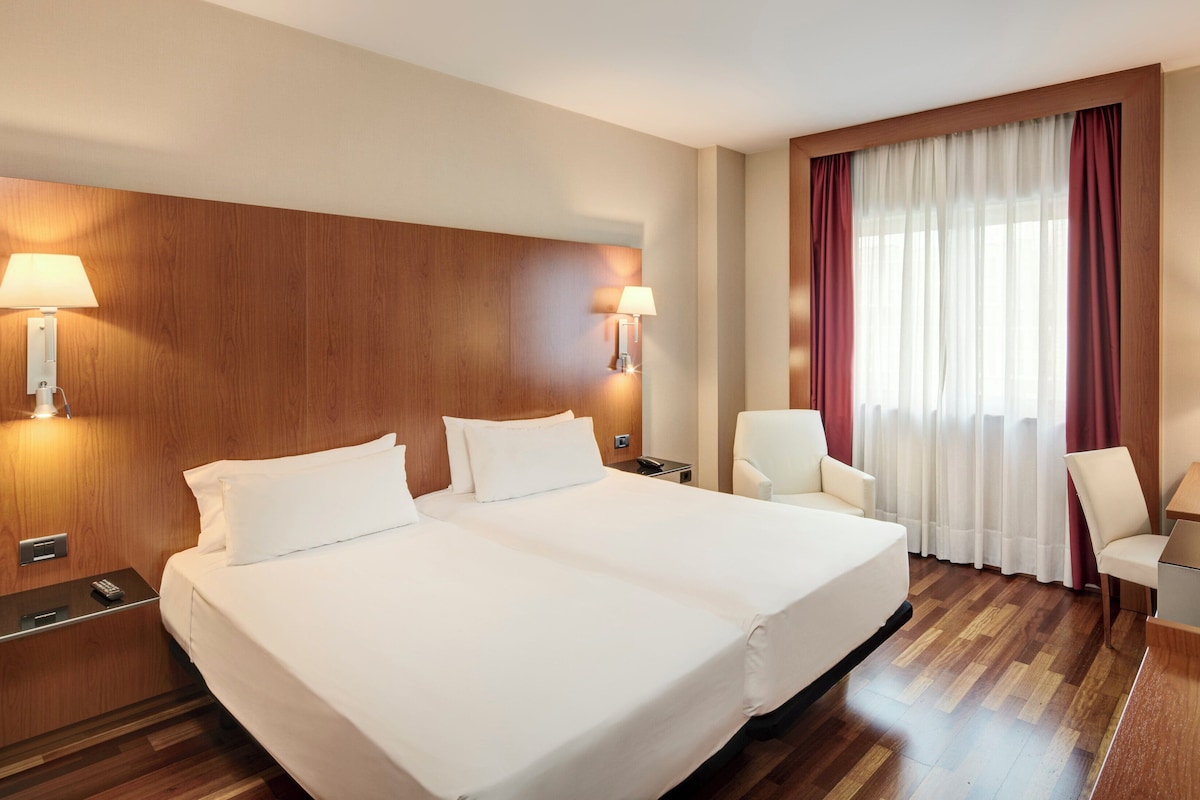 A hotel room with a large bed, two bedside tables, a desk with a chair, a TV, and a window with curtains.