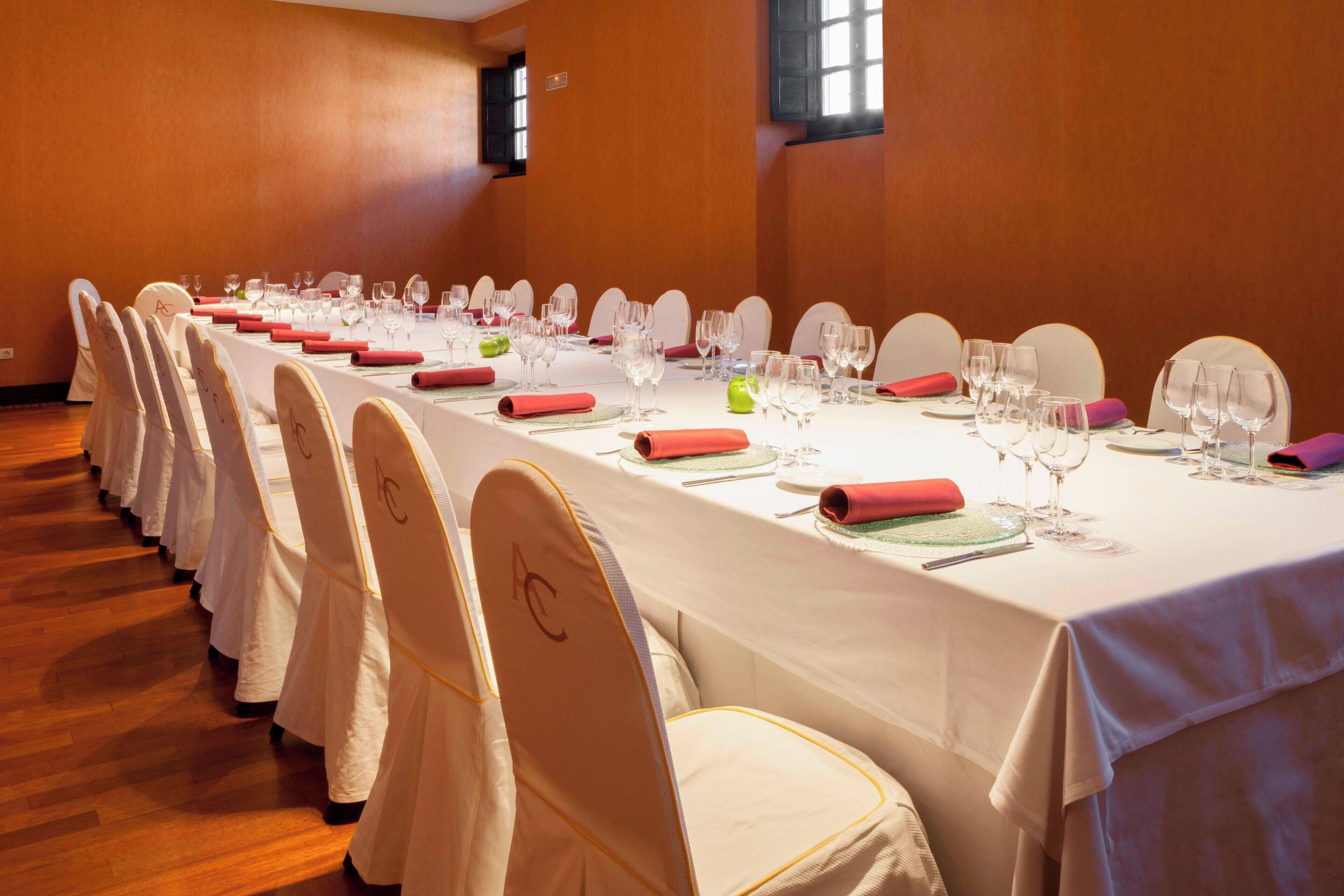 Una larga mesa de conferencias con manteles blancos, servilletas rojas y cristalería dispuesta para una reunión.