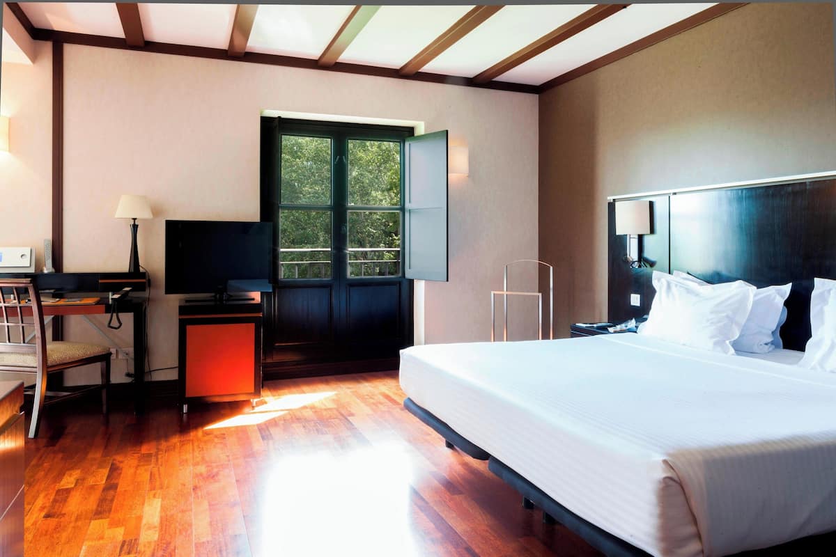 Quarto de hotel com cama, escrivaninha com computador, televisão e janela com vista para árvores.