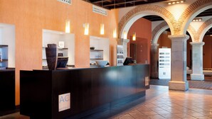 Eine Rezeption mit einem Schild, das "AC Hotels" zeigt, ein gefliestes Parkett und ein Backsteinbogen im Hintergrund.