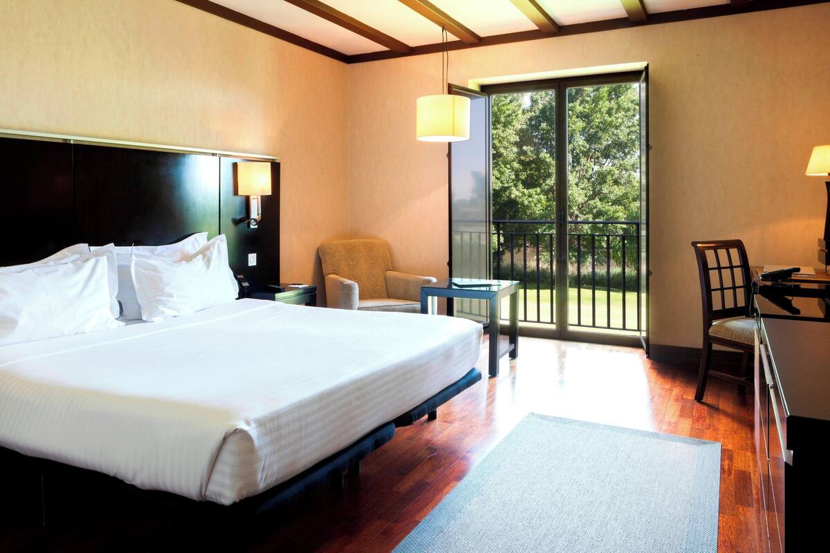 Quarto de hotel com uma cama grande, uma cadeira, uma mesa pequena e uma varanda com vista para árvores.