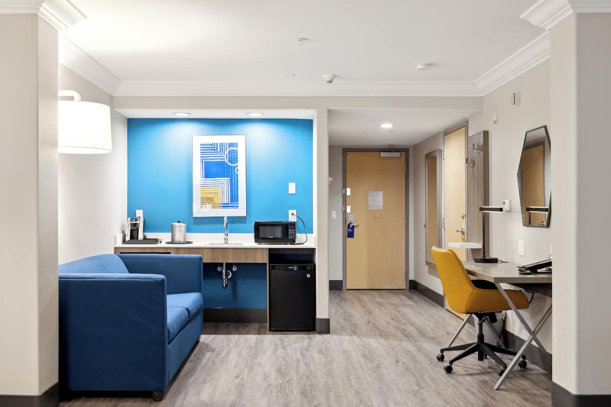 Ein modernes Hotelzimmer mit blauer Akzentwand, einer Küchenzeile, einem Sofa, einem Schreibtisch mit Stuhl und einem durchgehenden Türspalt sichtbaren Badezimmer.
