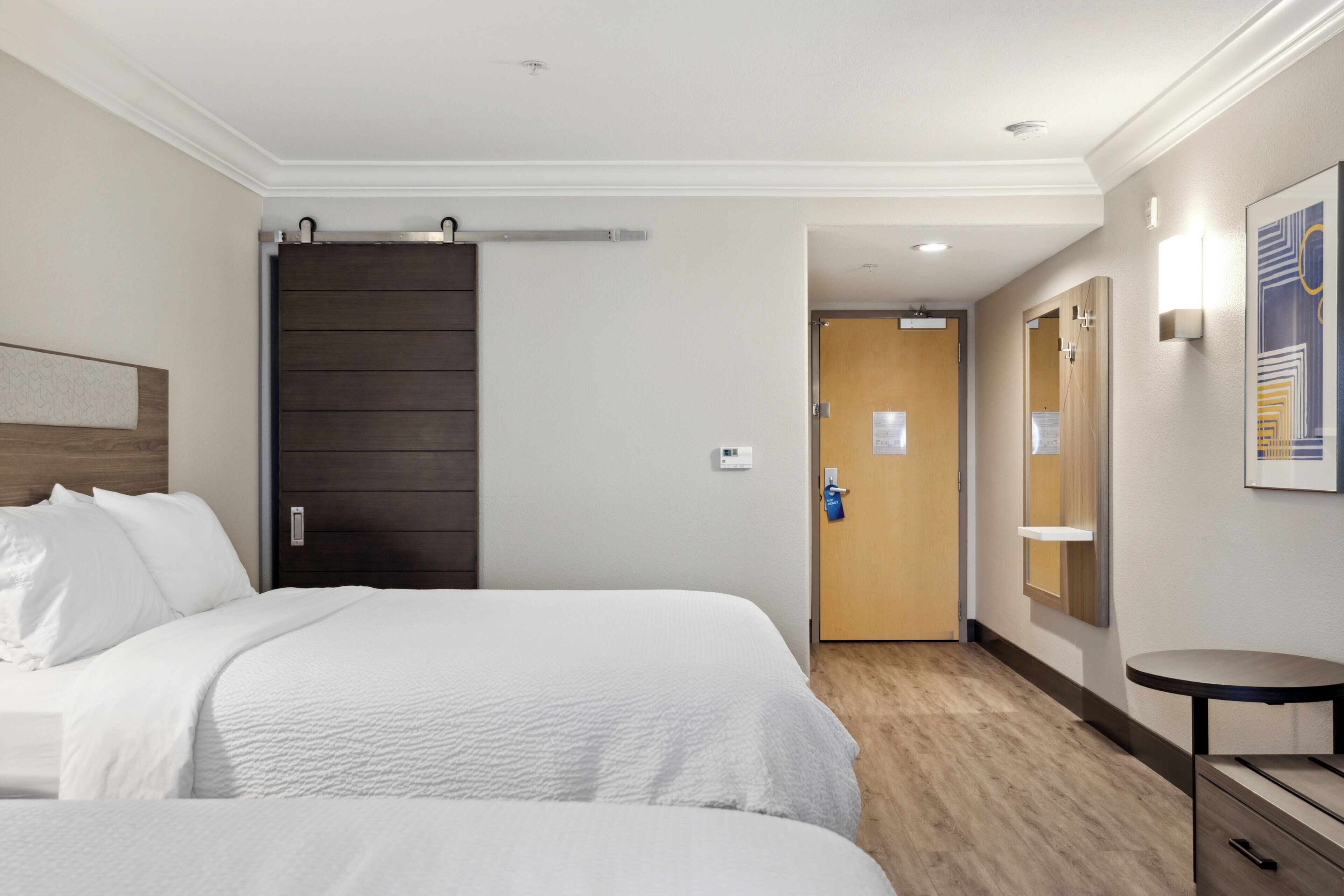 Una habitación de hotel con dos camas, una puerta de madera, una mesita y un aplique de pared.