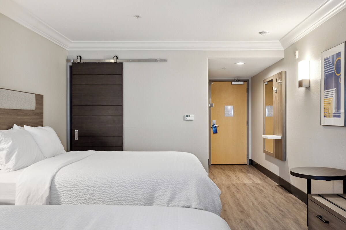A hotel room with two beds, a wooden door, a small table, and a wall-mounted light fixture.