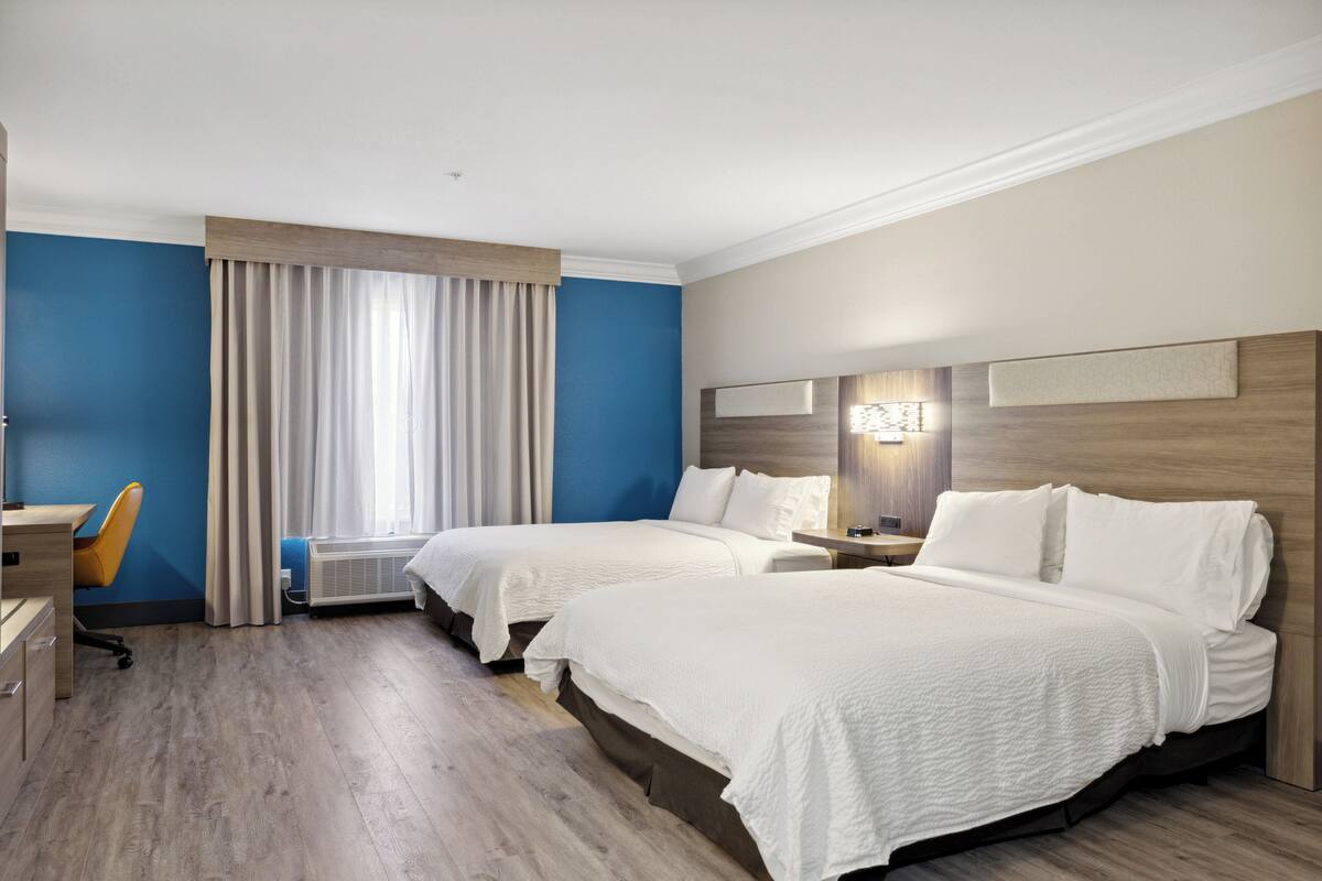 Ein Hotelzimmer mit zwei Betten, einem Schreibtisch, einem Stuhl und einem fensterbepflanzten Fenster.