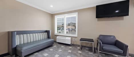 A room with a sofa, a chair, a flat-screen TV, a window with blinds, and a tiled floor.