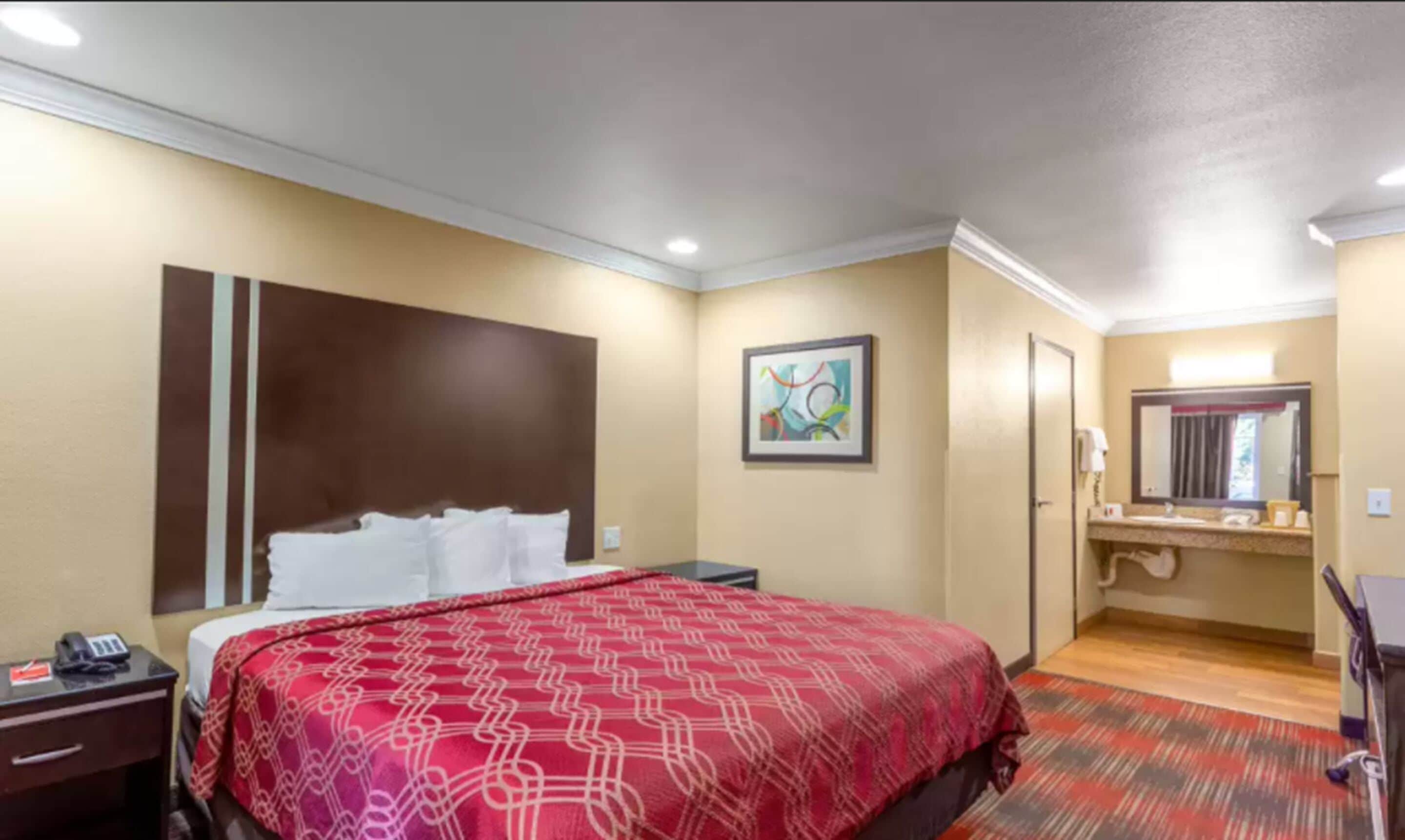 Ein Hotelzimmer mit einem großen Bett, einem Nachttisch mit Telefon, einem Bild an der Wand und einem Badezimmer mit Spiegel und Waschbecken.