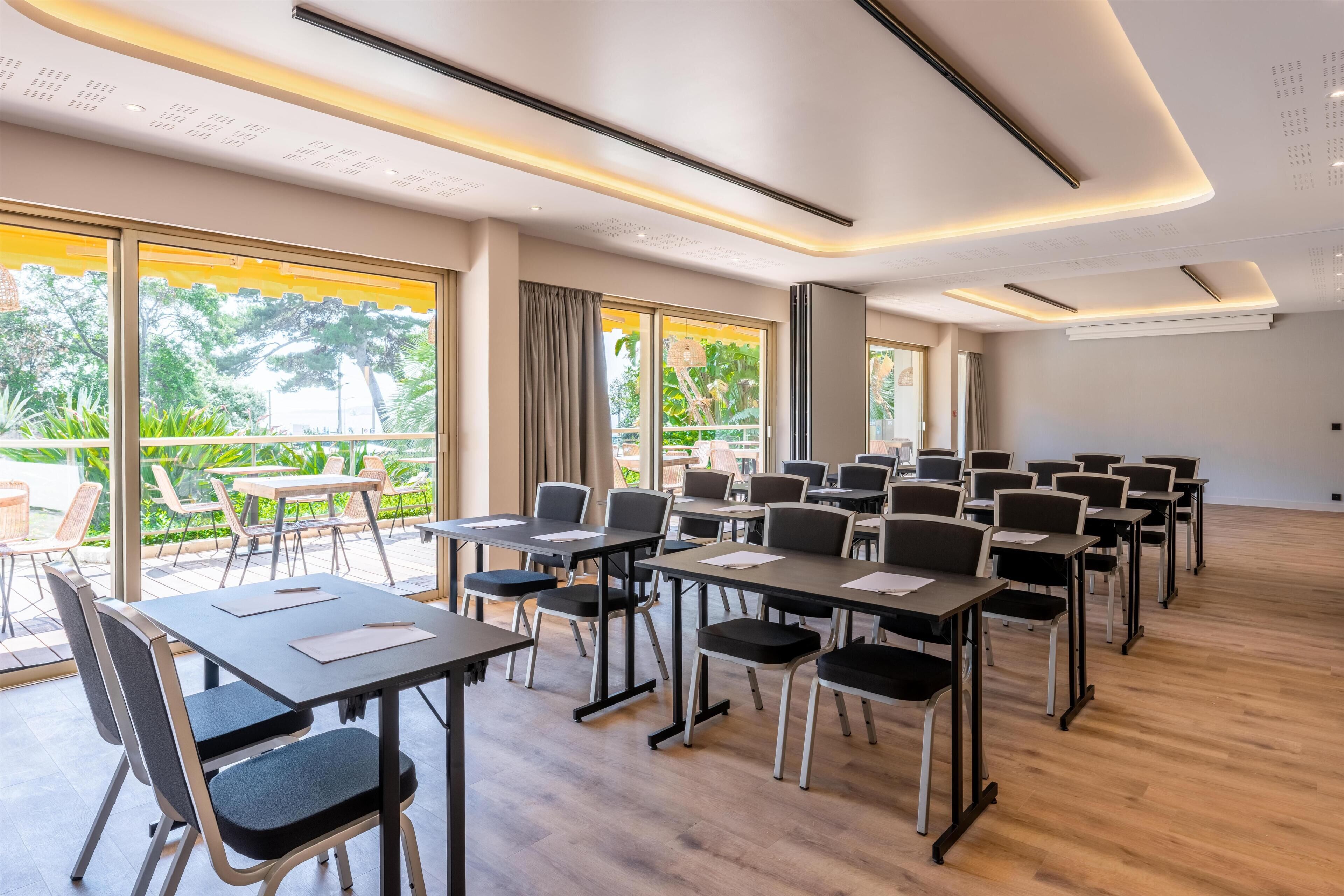Un salón de conferencias con mesas y sillas, ventanas amplias con cortinas y un balcón con mobiliario exterior.