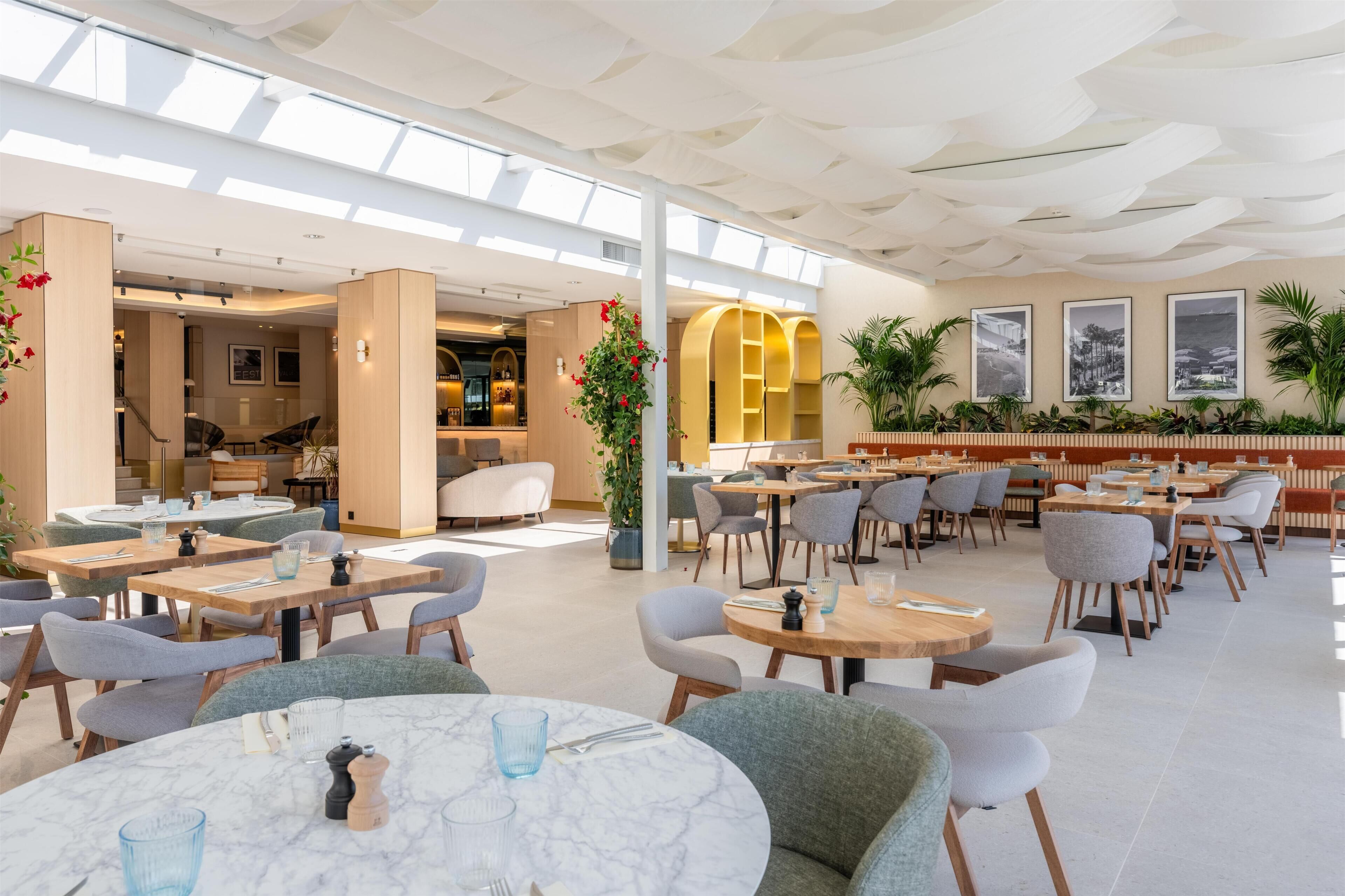 Een ruime binnenruimte met tafels en stoelen, een hoog plafond met een uniek ontwerp, en een zithoek met uitzicht.