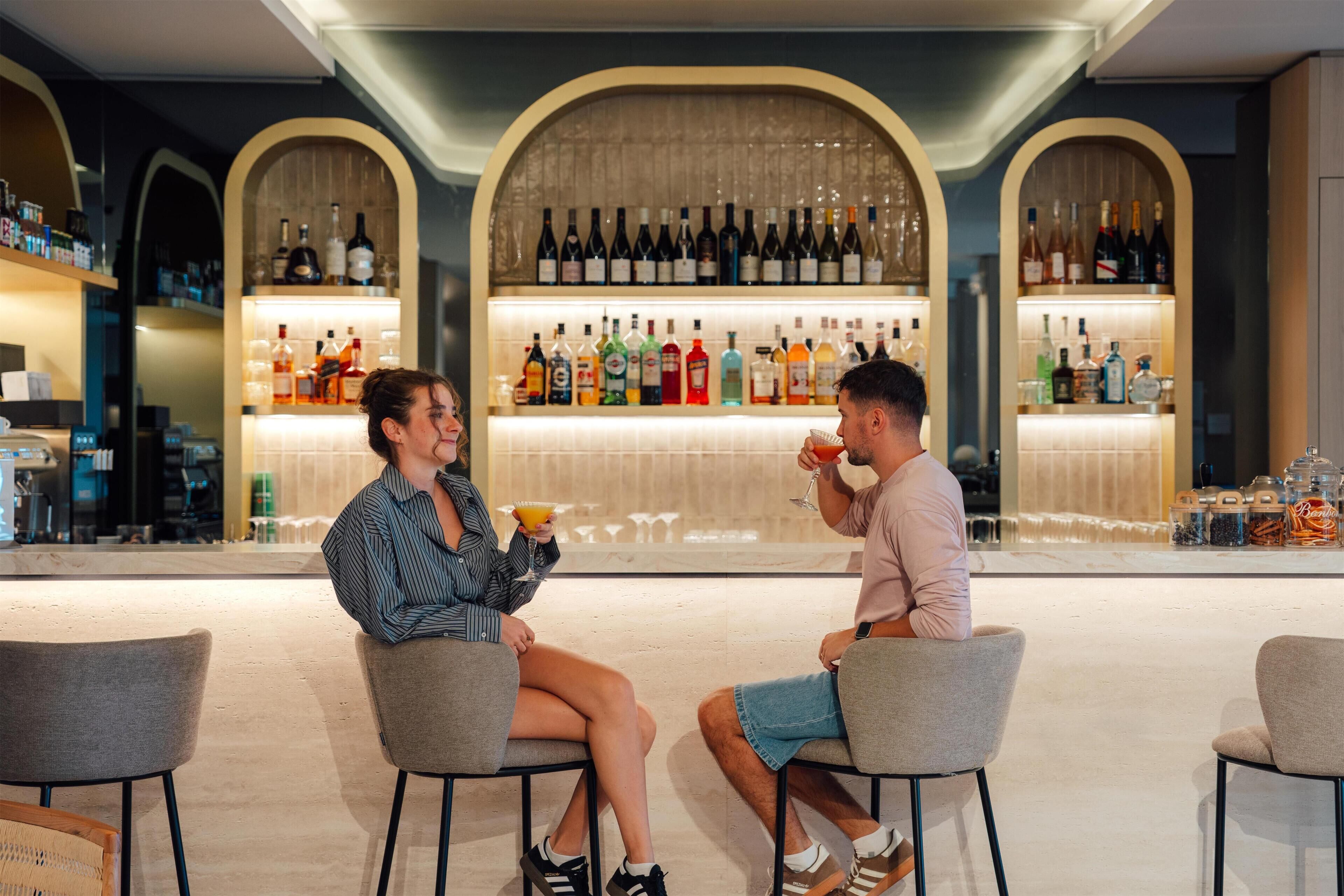 Two people seated at a bar with a variety of bottles and glasses in the background.