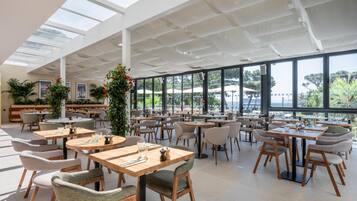 A spacious indoor dining area with multiple tables and chairs, large windows offering views of the outdoors, and a high ceiling with skylights.