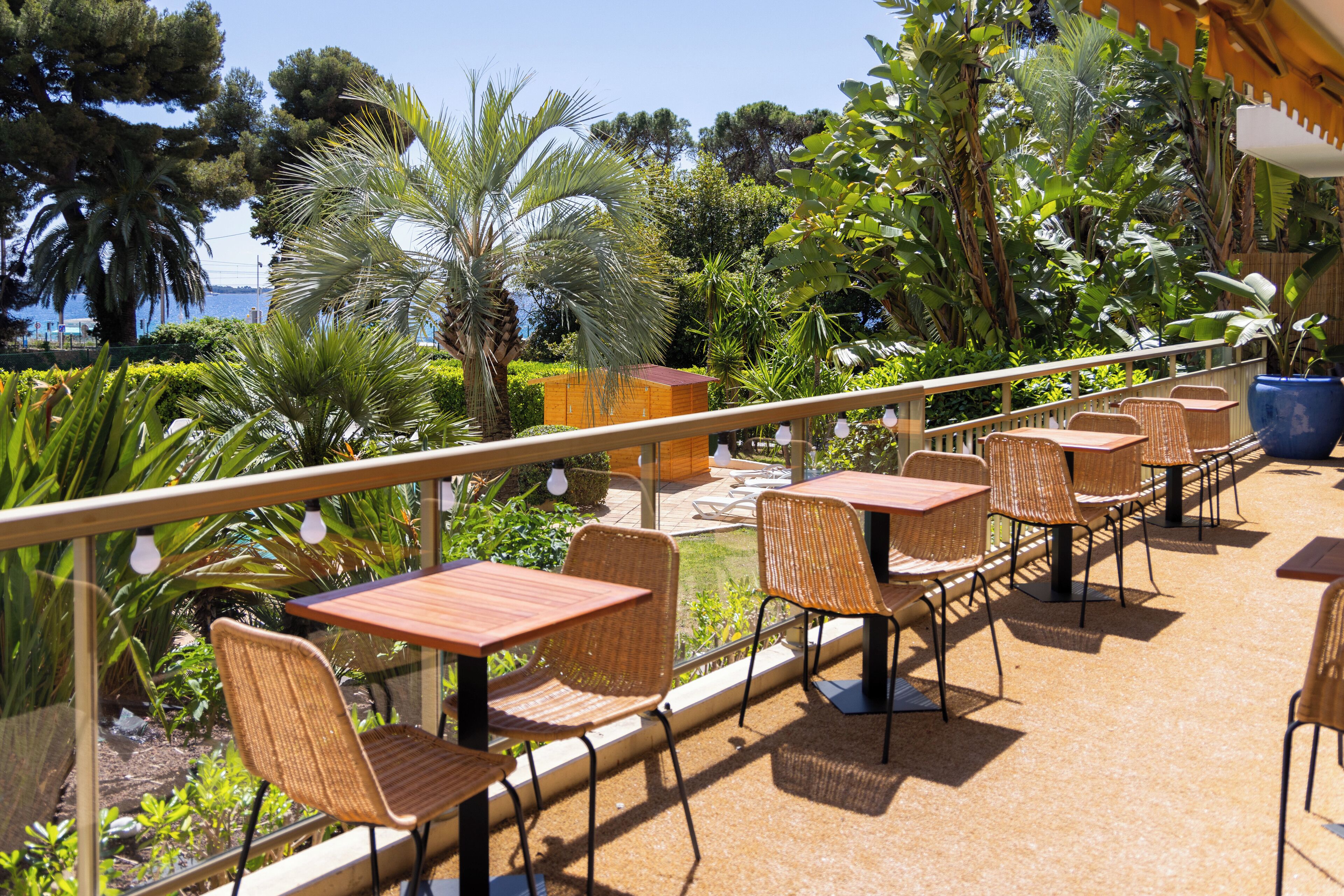 Un patio exterior con sillas y mesas de mimbre, rodeado de exuberante vegetación y vistas al mar.