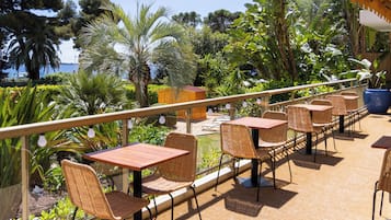 Un patio exterior con sillas y mesas de mimbre, rodeado de exuberante vegetación y vistas al mar.