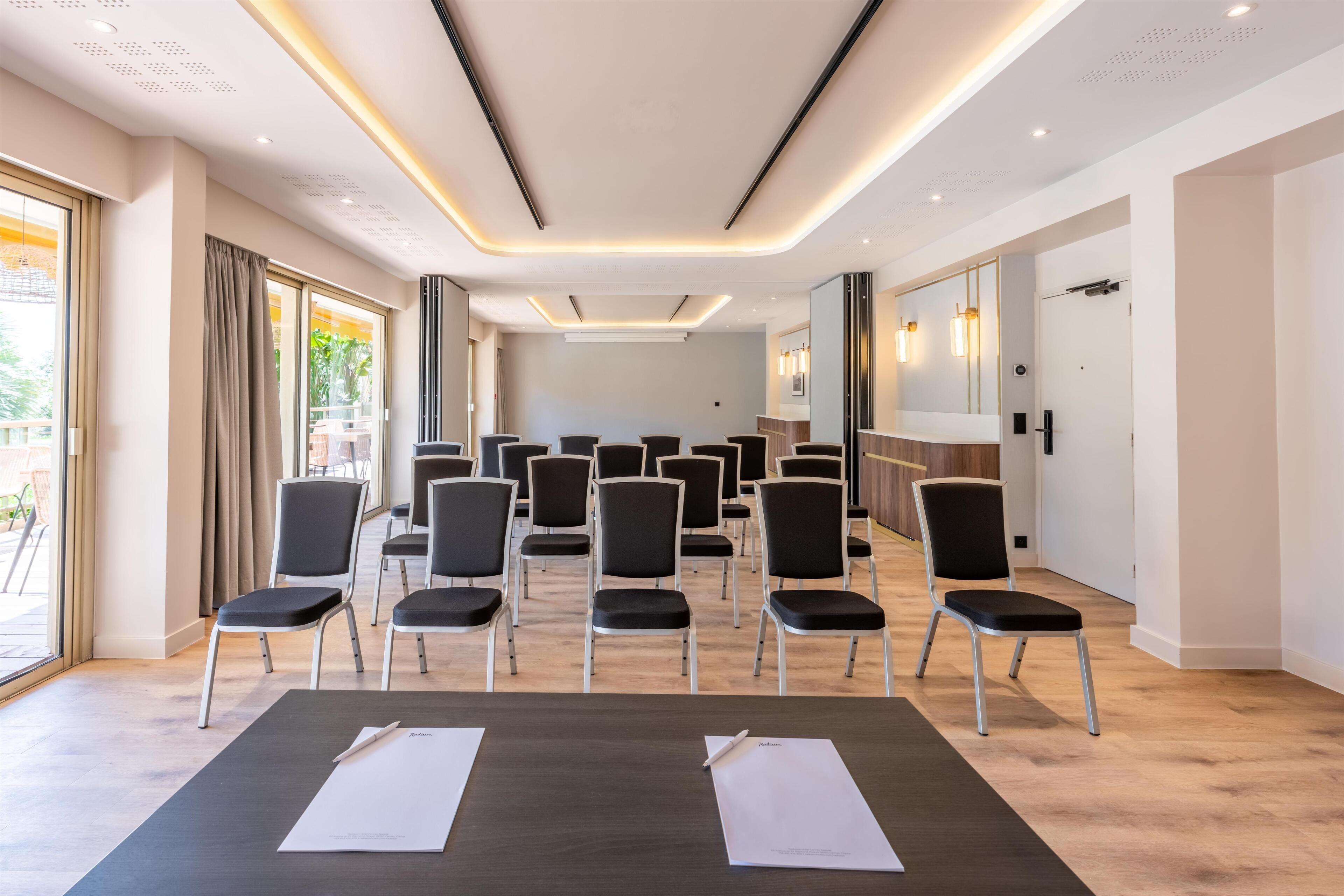 Un salón de conferencias con filas de sillas, una mesa con papeles y un espejo grande.