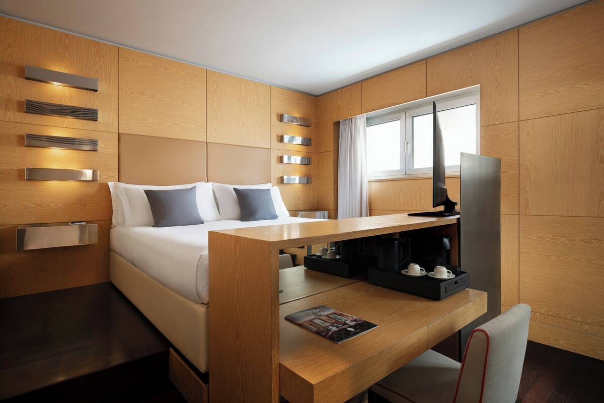 A modern hotel room with a bed, wooden panel walls, a desk with a coffee maker, and a window.