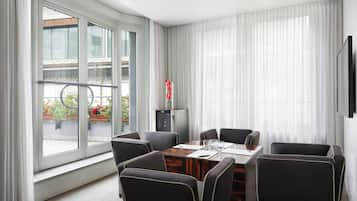 A modern hotel room with a large window, a dining table with chairs, a flat-screen TV, and a small cabinet.