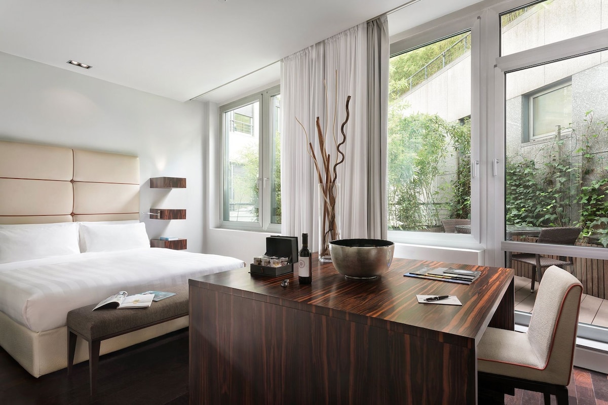 A modern hotel room with a bed, a wooden desk, a chair, and large windows offering a view of greenery.