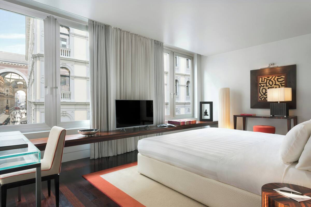 A hotel room with a large bed, a desk, a TV, and a view of a cityscape through the window.