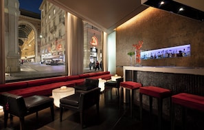 A bar area with seating, a bar counter with bottles, and a view of a street with shops. - Sina The Gray (Milan)
