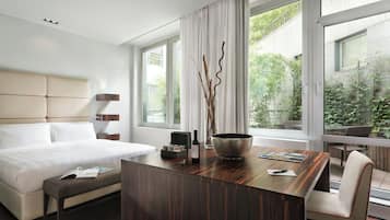 A modern hotel room with a large bed, a wooden desk, a chair, and a view of greenery outside.